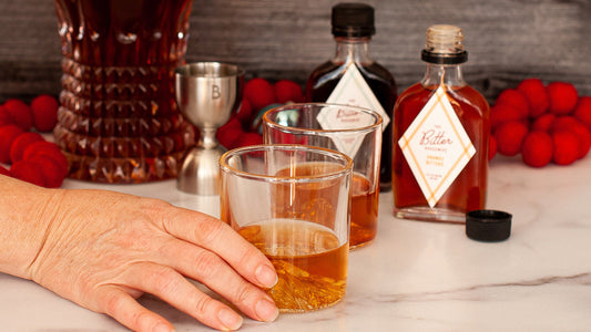 A hand reaching for an Old Fashioned with orange and aromatic bitters in the background.